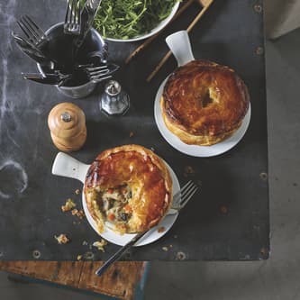 Two baked pies on a table ready to eat with cutlery, salt, pepper, and a green salad